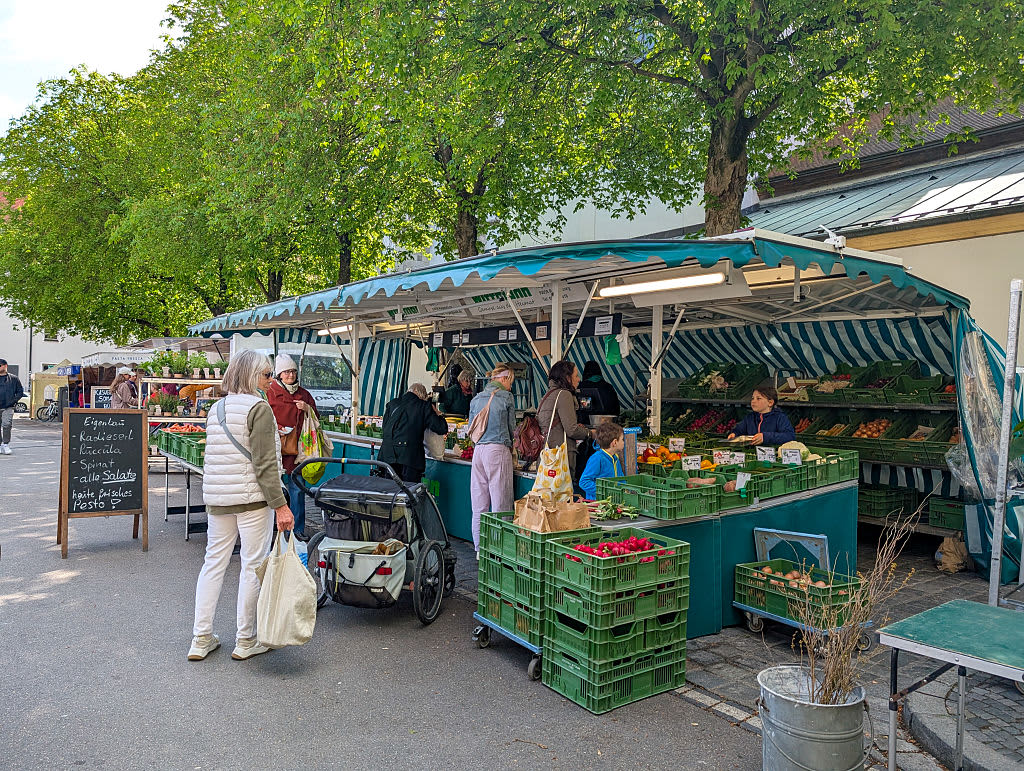 Shoppers buy fresh vegetables, fruit, and herbs at an outdoor produce market under green-striped canopies in Regensburg, Upper Palatinate, Bavaria, Germany, on April 19, 2025.